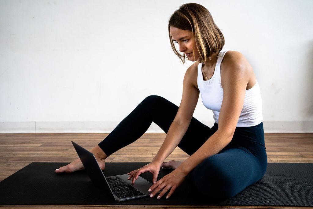 A woman sitting cross legged on a mat beside an open laptop for a live online Pilates class