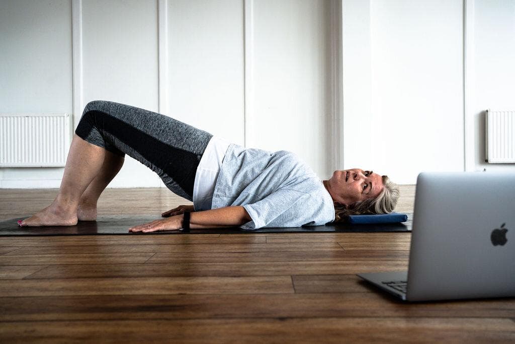 A member holding a shoulder bridge on a mat while following a recorded class on a laptop
